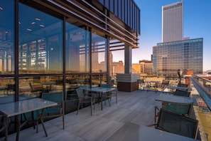 An outdoor seating area with tables and chairs, large windows, and a view of a city skyline. - Hotel Indigo Tulsa DWTN-Entertainment Area by IHG (Tulsa)