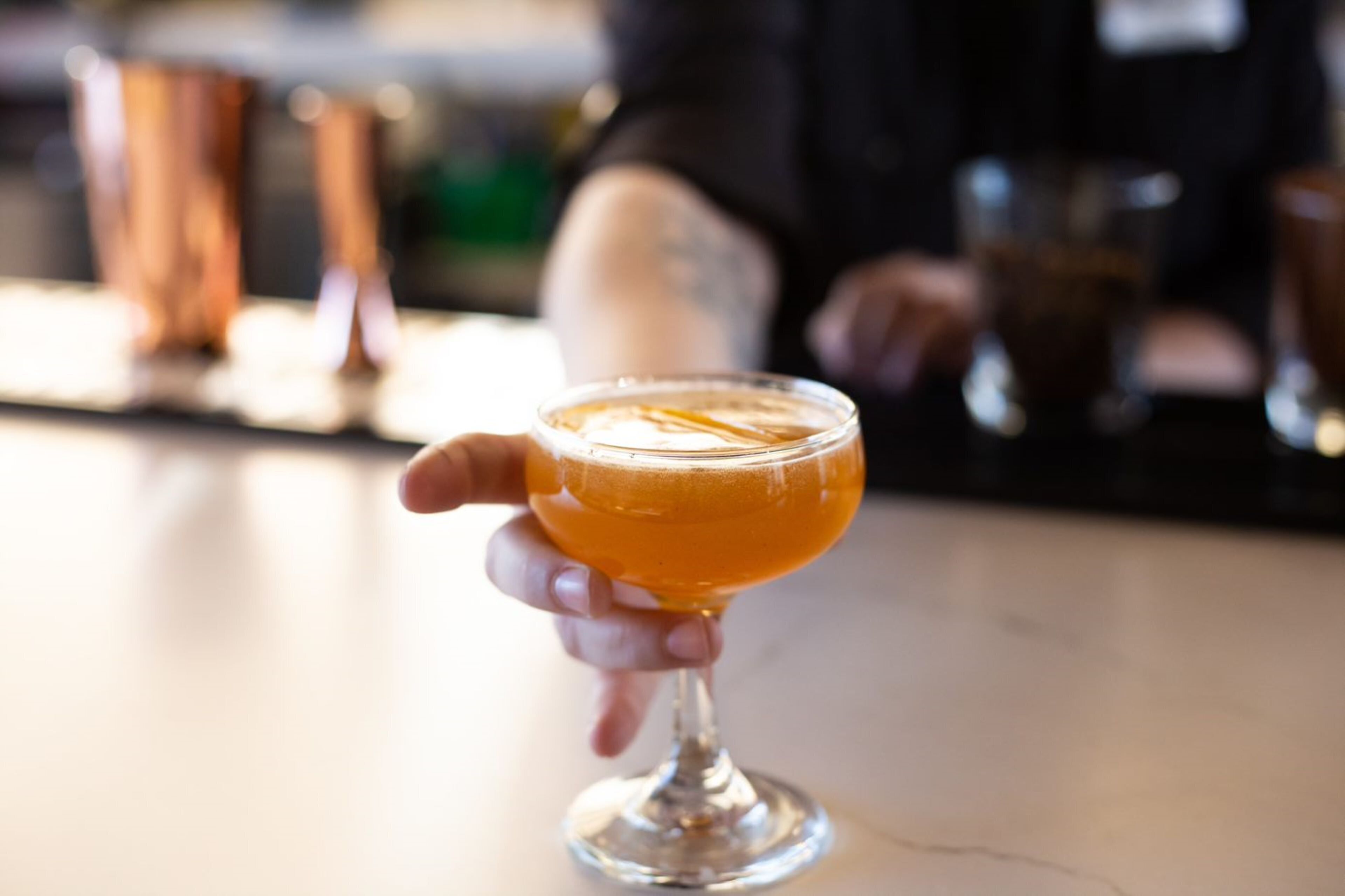 A hand holding a cocktail glass with a golden liquid, possibly a whiskey-based drink, on a bar counter.