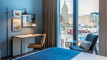Une chambre d’hôtel avec un grand lit, un bureau, une chaise et une vue sur le paysage urbain grâce à de grandes fenêtres.