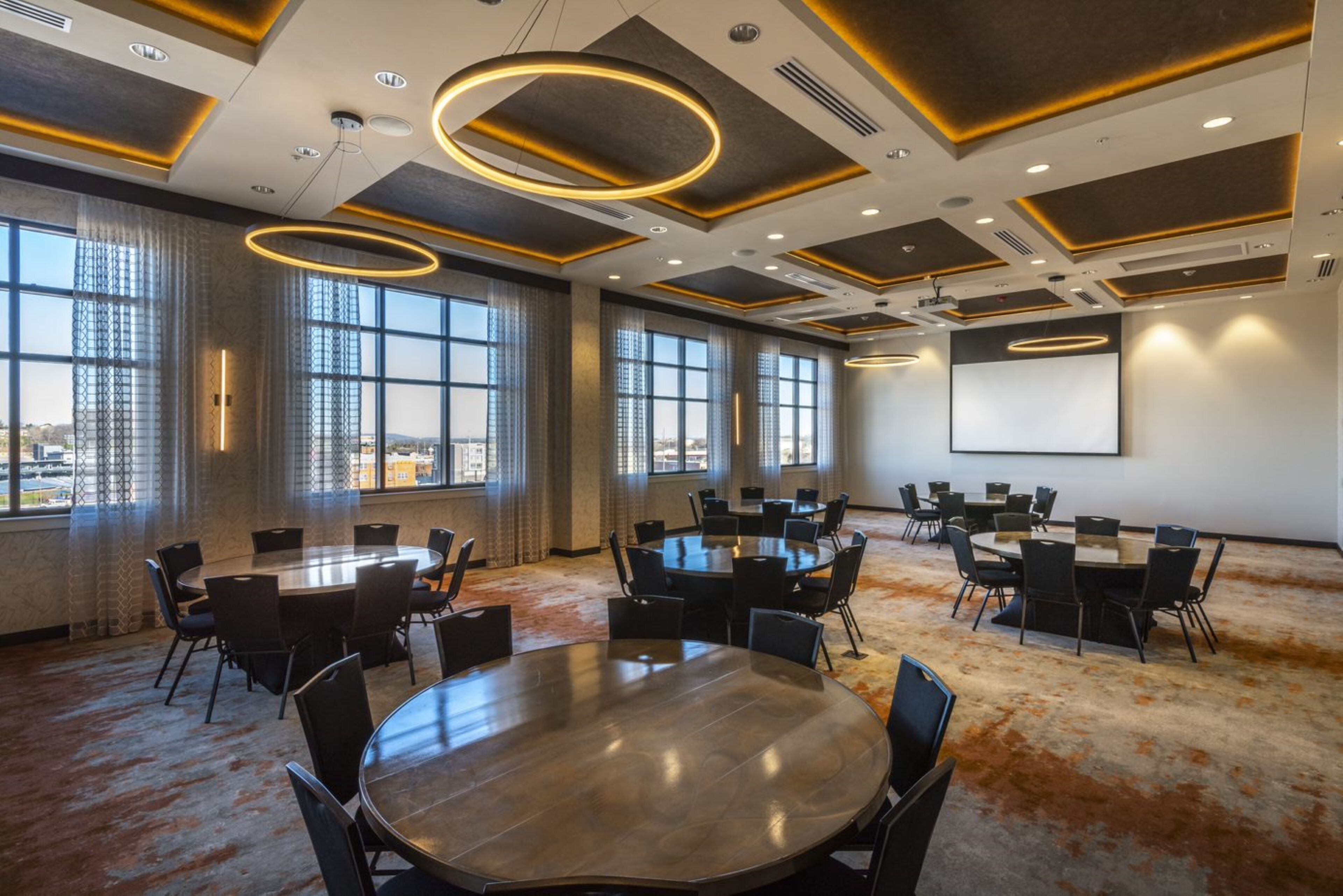 A conference room with multiple tables and chairs, large windows, and a projector screen.