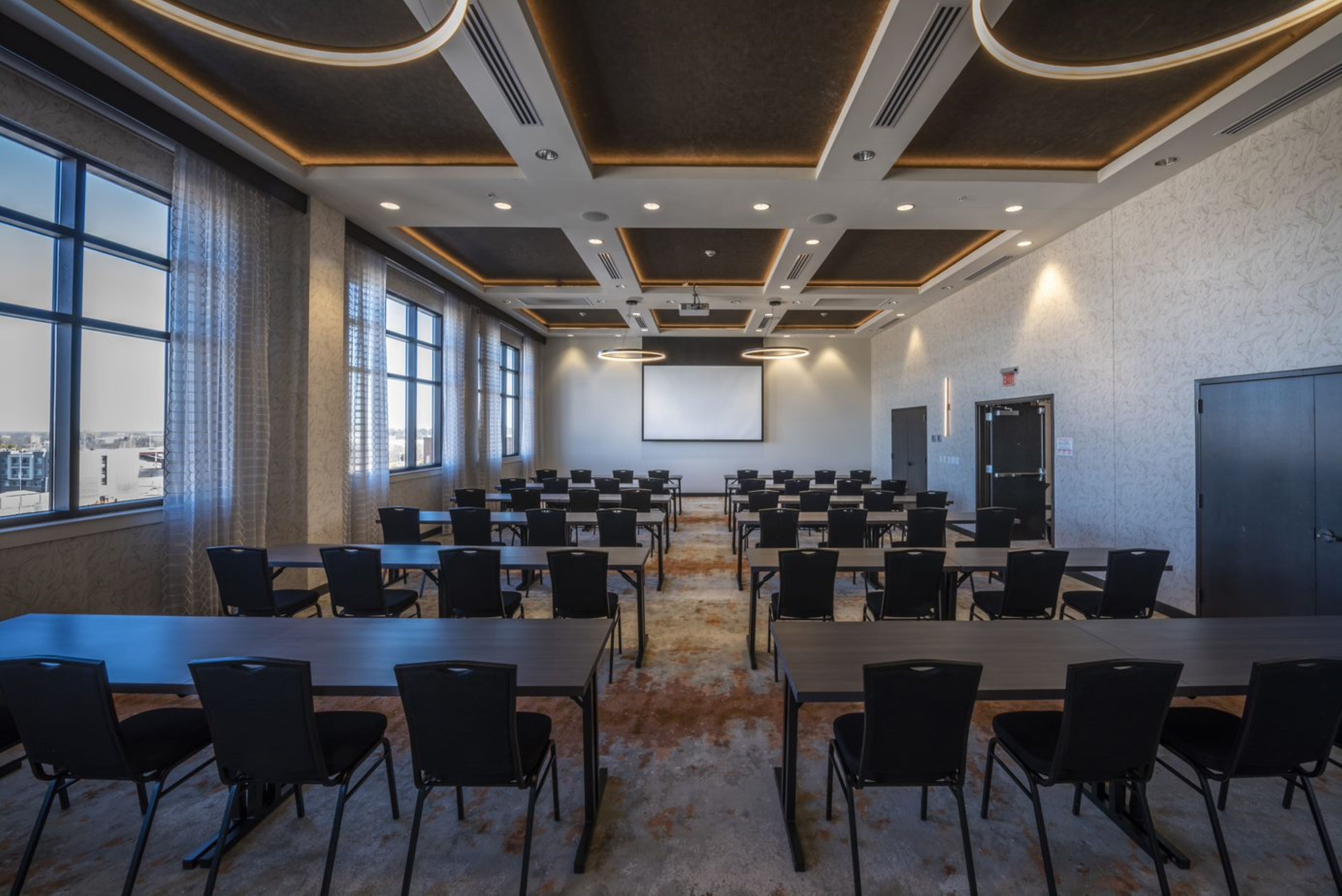 A conference room with multiple tables and chairs, large windows, and a projector screen.