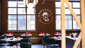Interior de restaurante com paredes de tijolo, grandes janelas e um logotipo central. Mesas com almofadas vermelhas e copos.