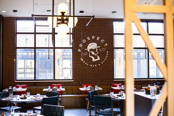 A restaurant interior with brick walls, large windows, and a central logo. Tables are set with red cushions and glasses. - Hotel Indigo Tulsa DWTN-Entertainment Area by IHG (Tulsa)