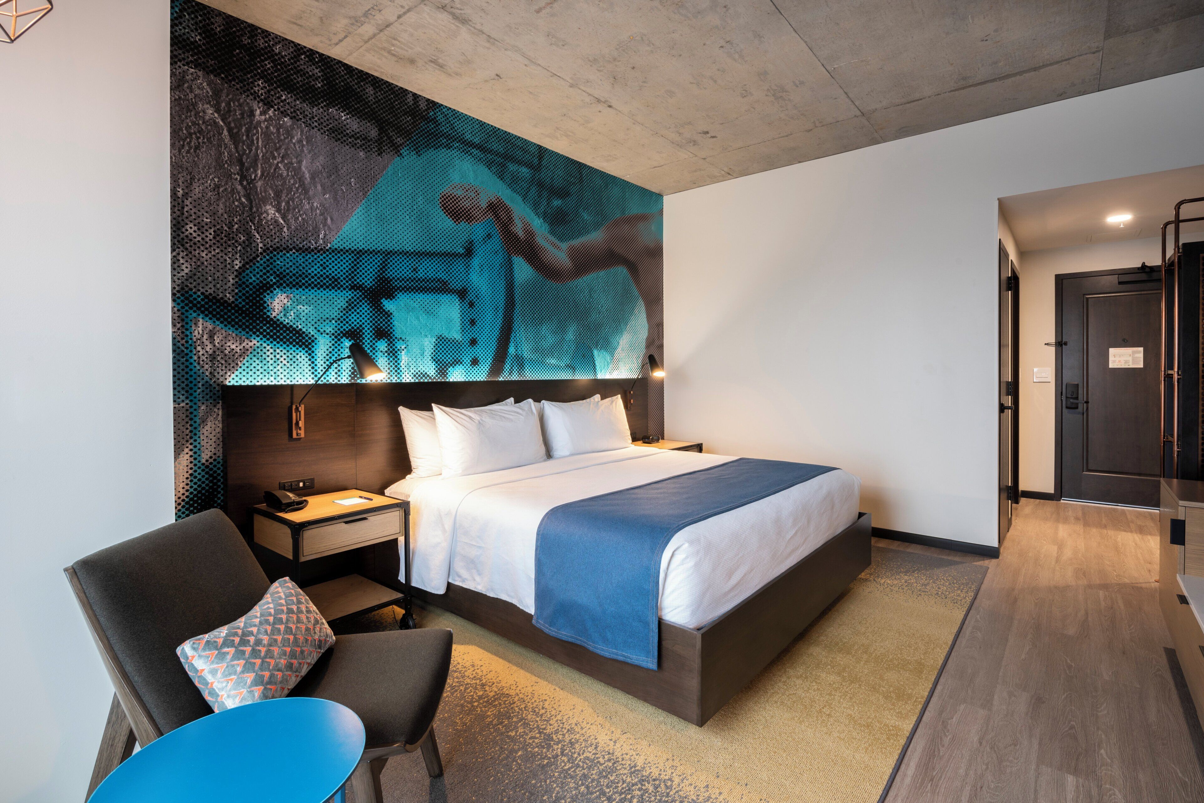 A modern hotel room with a large bed, a desk, a chair, and a wall-mounted artwork.