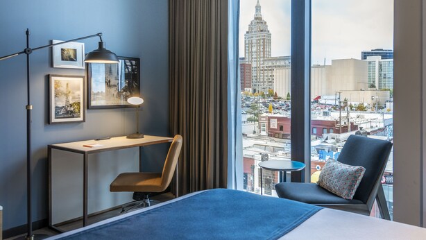Quarto de hotel com uma cama grande, uma escrivaninha, uma cadeira, uma luminária e vista para a cidade através de janelas amplas.