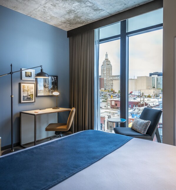 A hotel room with a large bed, a desk, a chair, a lamp, and a view of a cityscape through large windows. - Hotel Indigo Tulsa DWTN-Entertainment Area by IHG (Tulsa)