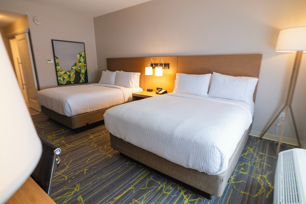 A hotel room with two beds, a headboard with integrated lighting, a bedside table with a phone, and a carpeted floor.