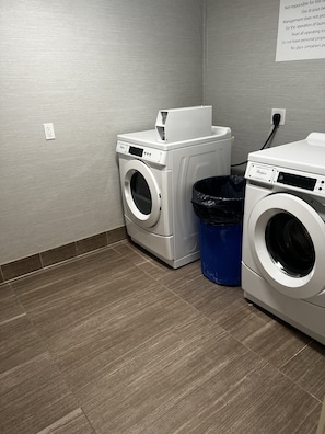 A laundry room with two white front-loading washing machines, a blue trash bin, and a wall-mounted electrical outlet.