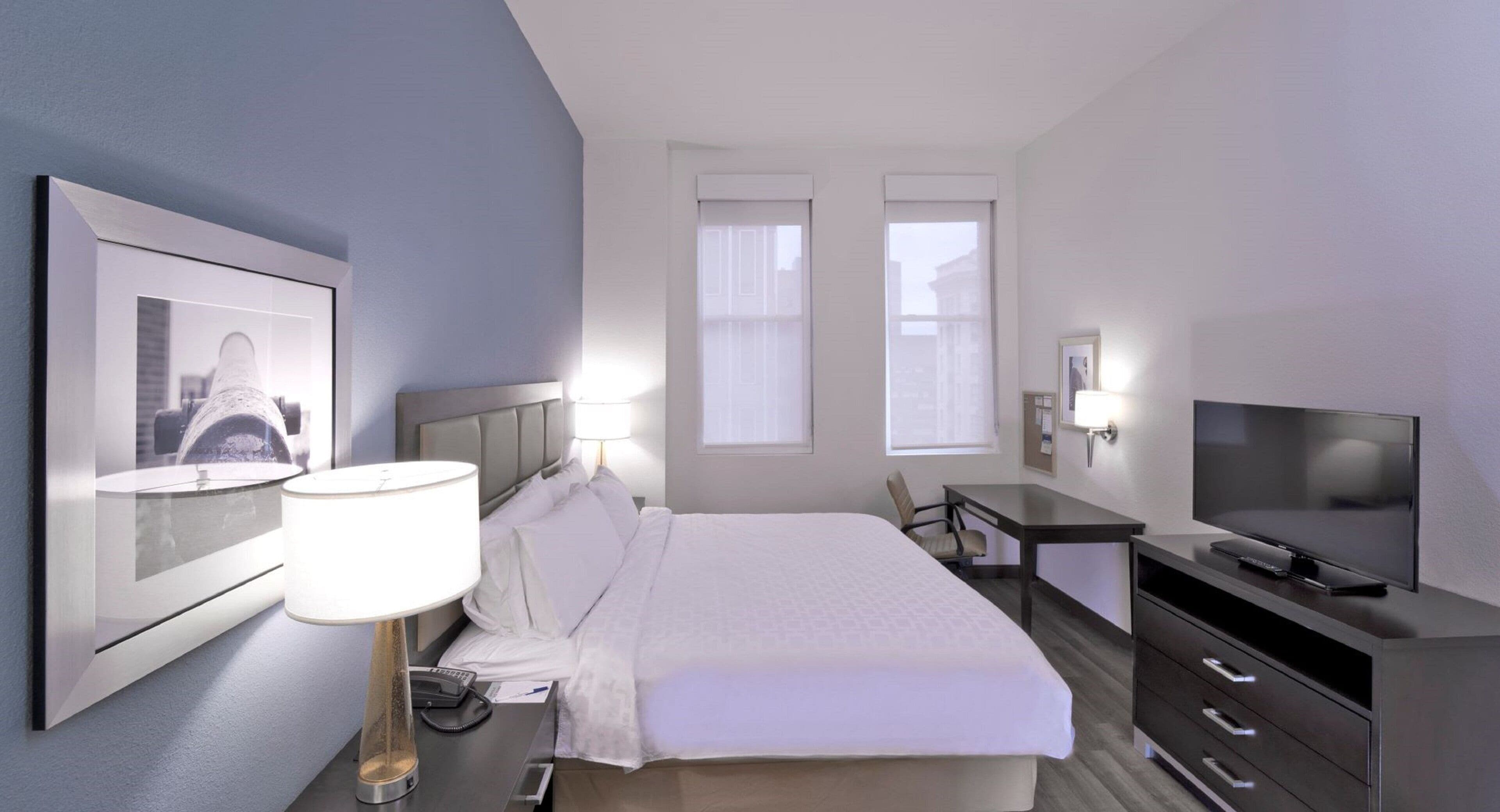 A hotel room with a bed, bedside table, lamp, TV, and a framed picture on the wall.