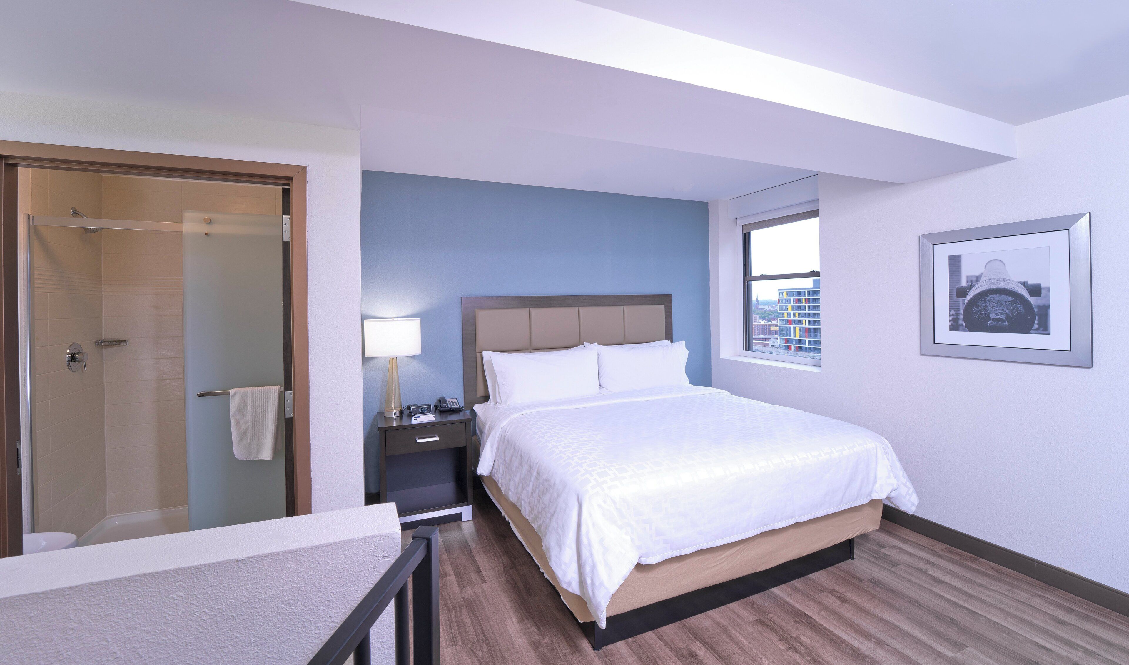 A hotel room with a bed, a bedside table, a lamp, a window with a city view, and a door leading to a bathroom.