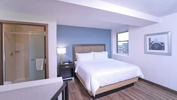 A hotel room with a bed, a bedside table, a lamp, a window with a city view, and a door leading to a bathroom.