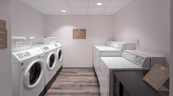 Un cuarto de lavandería con varias lavadoras y secadoras, un cesto de cortesía y un letrero que indica lavandería gratuita para huéspedes.