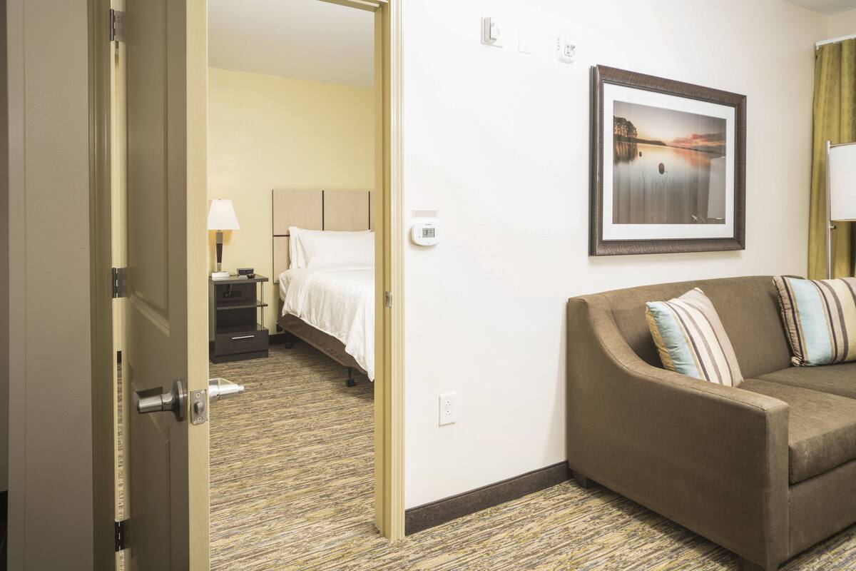 A hotel room with a bed, a brown sofa, a framed picture on the wall, and a carpeted floor.