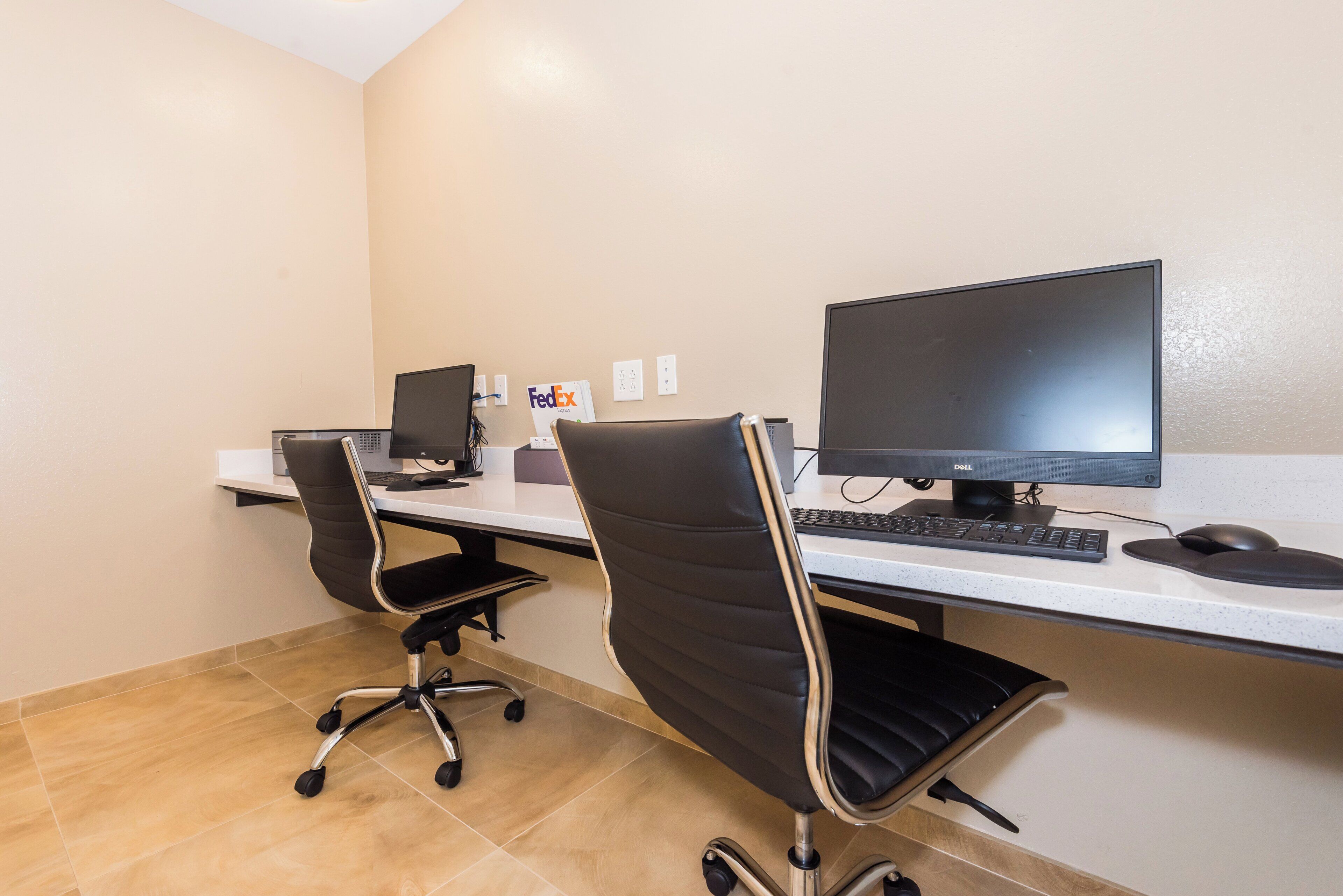 A workspace with two desks, each with a computer, keyboard, and mouse. There are two office chairs, one with wheels and one without. The room has a tiled floor and a plain wall with a few electrical outlets.
