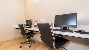 Un espacio de trabajo con dos escritorios, cada uno con una computadora, teclado y ratón. Hay dos sillas de oficina, una con ruedas y otra sin. El cuarto tiene piso de baldosas y una pared lisa con algunos tomacorrientes.