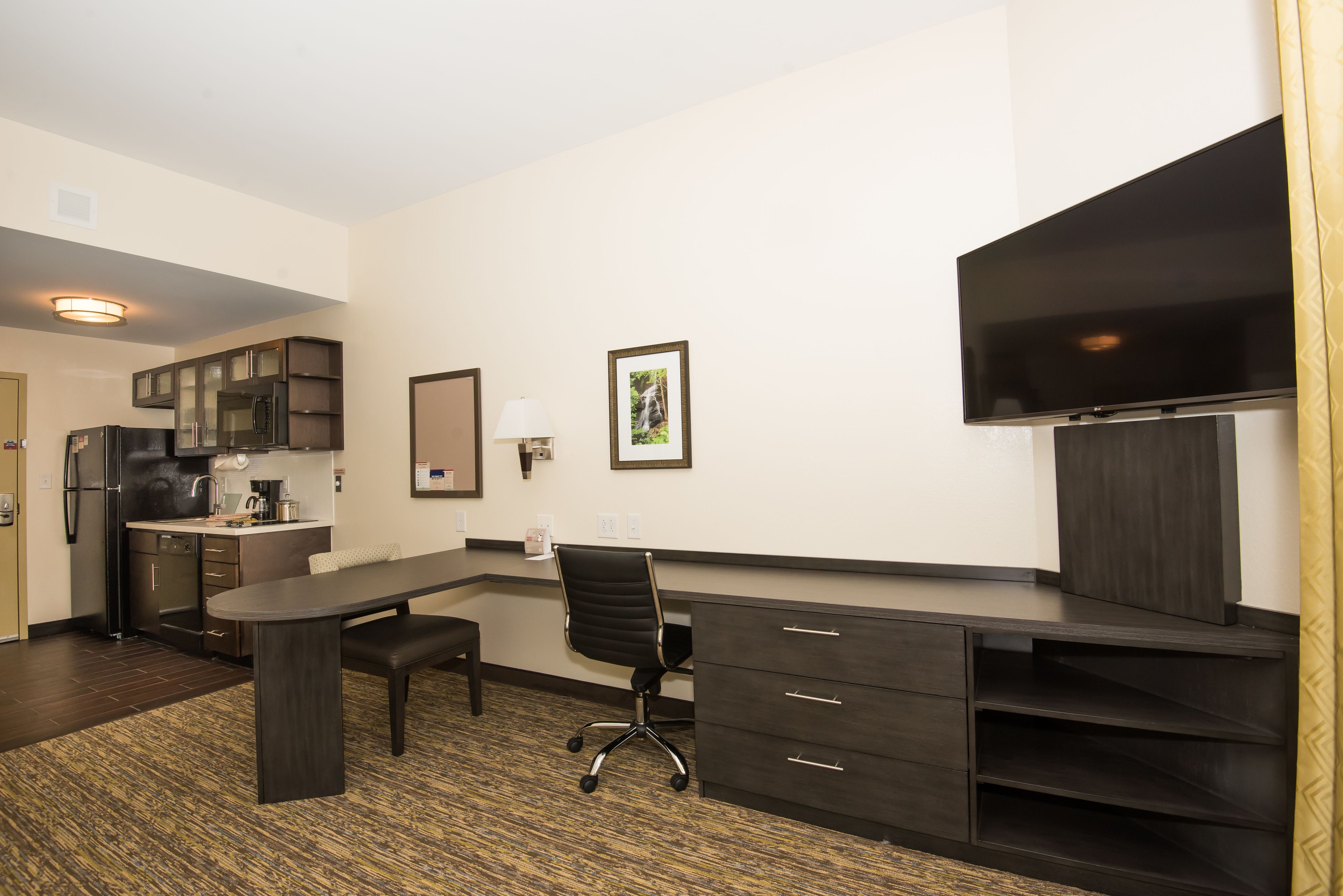 A modern hotel room with a kitchenette, a desk with a chair, a flat-screen TV, and a large window.