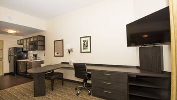 A modern hotel room with a kitchenette, a desk with a chair, a flat-screen TV, and a large window.