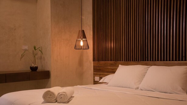 Una cama bien hecha con sábanas blancas y toallas enrolladas, un cabecero de madera, una lámpara colgante y una pequeña planta en maceta sobre una mesita de noche.