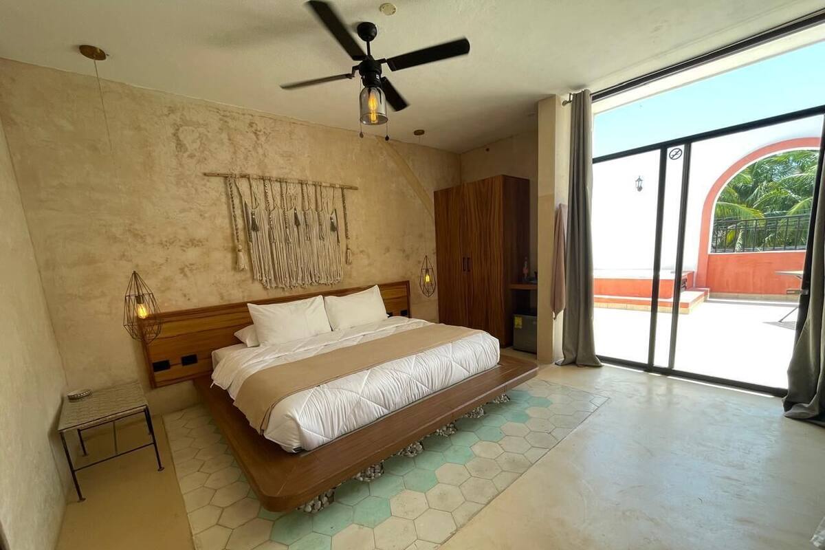 Ein Schlafzimmer mit einem großen Bett, einem Deckenventilator, einer Wandleuchte und Blick durch eine Glastür.