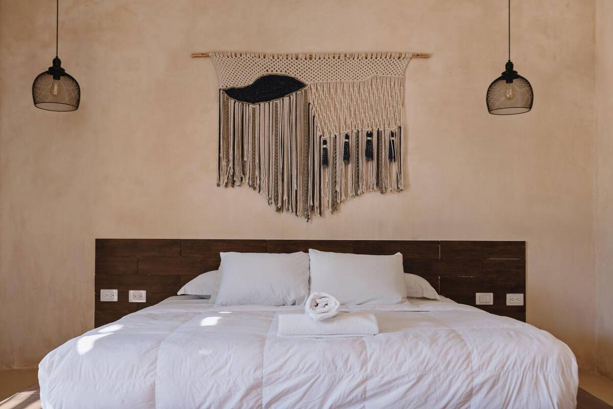 Ein ordentlich bezogenes Bett mit weißen Leinen, einem dunklen Holz-Kopfteil und einem Wandbehang aus Makramee darüber.