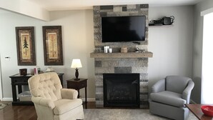 A living room with a stone fireplace, a flat-screen TV, two framed artworks, a beige armchair, a gray armchair, and a small table with a bowl.