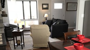 A living room with a sofa, a chair, a dining table with red placemats and a bowl, and a kitchen area with a lamp.