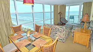 A room with a dining table, chairs, a sofa, a lamp, and a view of the ocean. - Oceanfront  Seaside Resort 1403 3BR/3BA. Resort Amenities with Amazing views (NO. MYRTLE BEACH)