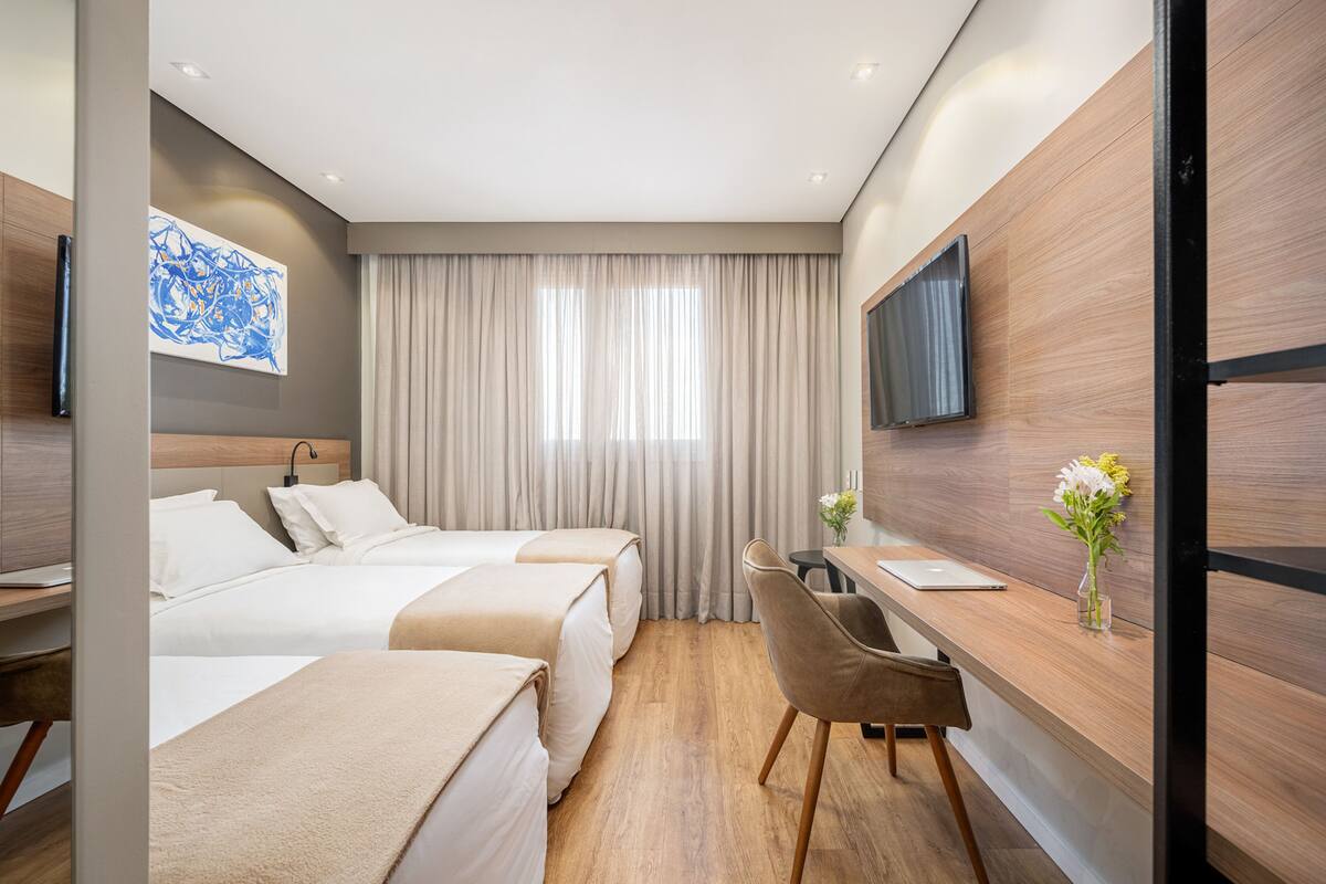 Ein Hotelzimmer mit zwei Betten, einem Schreibtisch, einem Stuhl, einem Fernseher und einem Fenster mit Vorhängen.
