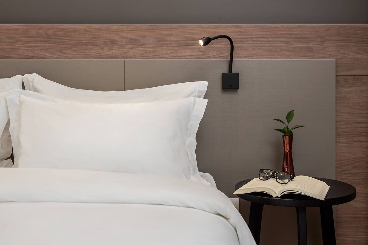 Ein ordentlich bezogenes Bett mit weißen Leinen, ein moderner Nachttisch mit einem Buch und Brille sowie zwei gerahmte Kunstwerke an der Wand.
