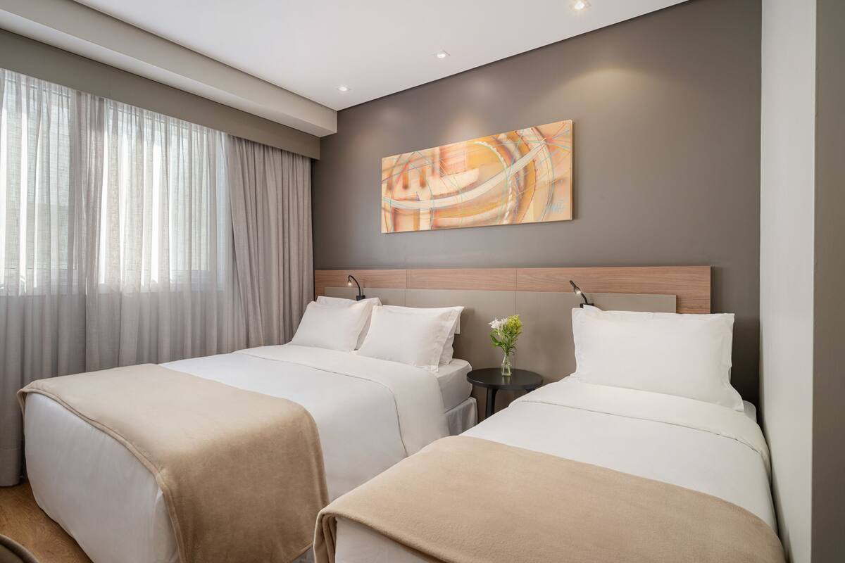 Ein Hotelzimmer mit zwei Betten, einem großen Fenster mit durchsichtigen Vorhängen, einem modernen abstrakten Gemälde an der Wand und einem kleinen Tisch mit einem Blumenstrauß.