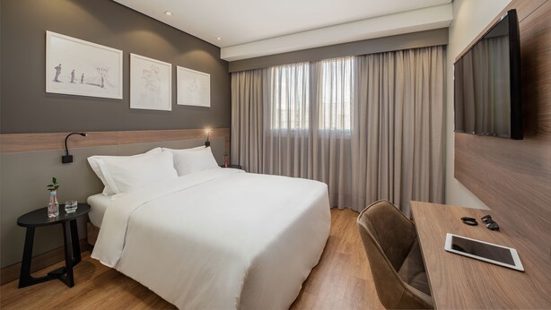 Ein Hotelzimmer mit Bett, Nachttisch mit Flasche und Gläsern, Schreibtisch mit Tablet und Sonnenbrille, Fernseher und einem Kunstwerk an der Wand.