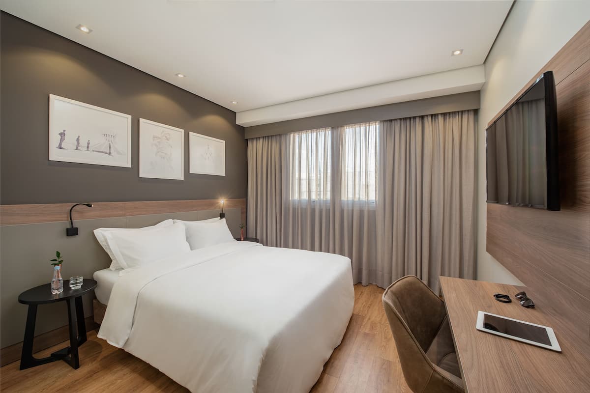Ein modernes Hotelzimmer mit einem ordentlich bezogenen Bett, einem Nachttisch mit Flasche und Gläsern, einem Schreibtisch mit Tablet und Sonnenbrille sowie einem großen Fernseher an der Wand.