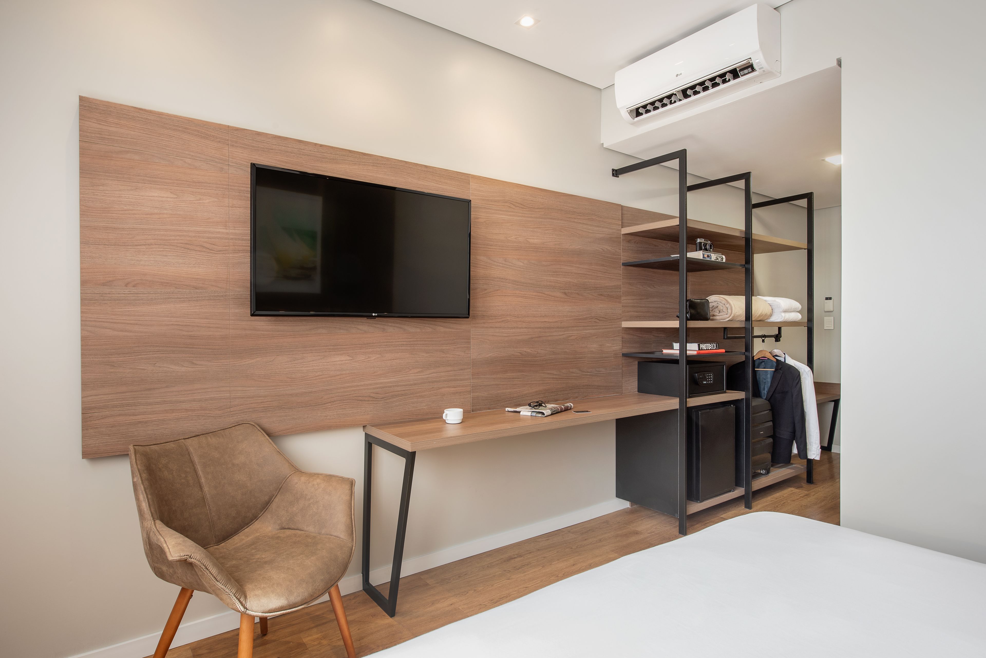 Quarto moderno com televisão na parede, escrivaninha de madeira, uma cadeira e roupeiro embutido com prateleiras.