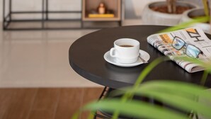 Gemütliche Innenraumansicht mit rundem Tisch, einer Kaffeetasse, einer Topfpflanze und einem Bücherregal im Hintergrund.