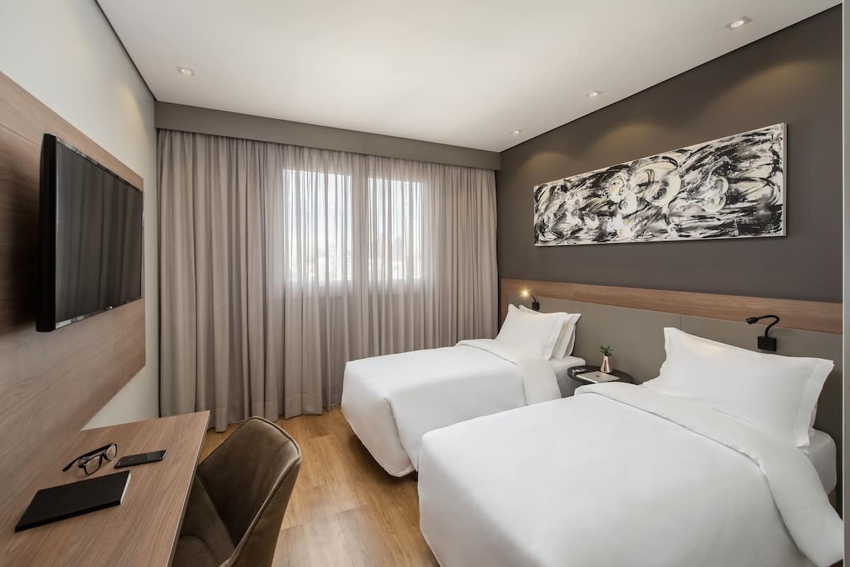 Ein Hotelzimmer mit zwei Betten, einem Schreibtisch mit Stuhl, einem Fernseher und einem großen abstrakten Gemälde an der Wand.