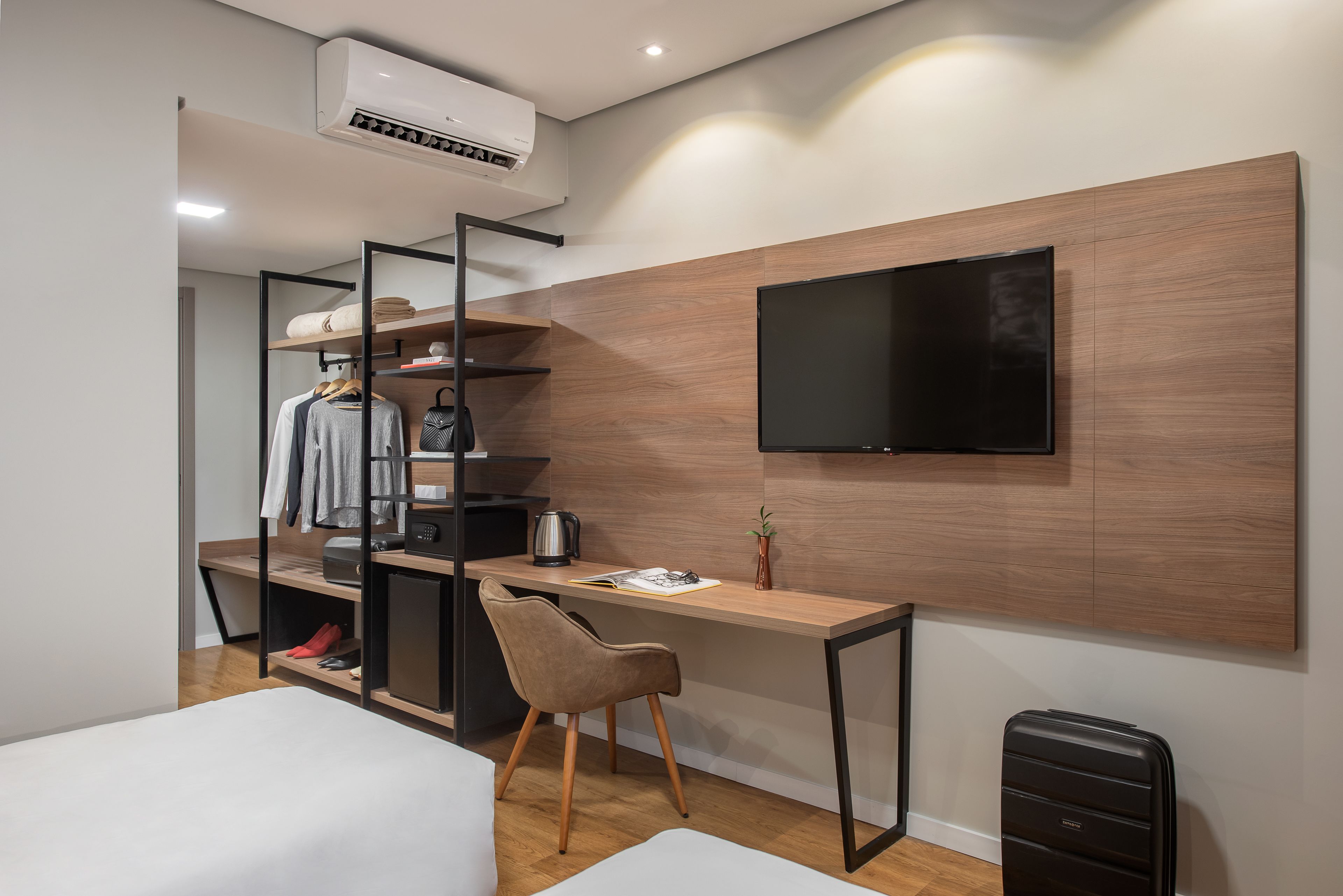 Una habitación de hotel moderna con un escritorio de madera, un televisor de pantalla plana en la pared, un armario con ropa colgada y una cama.