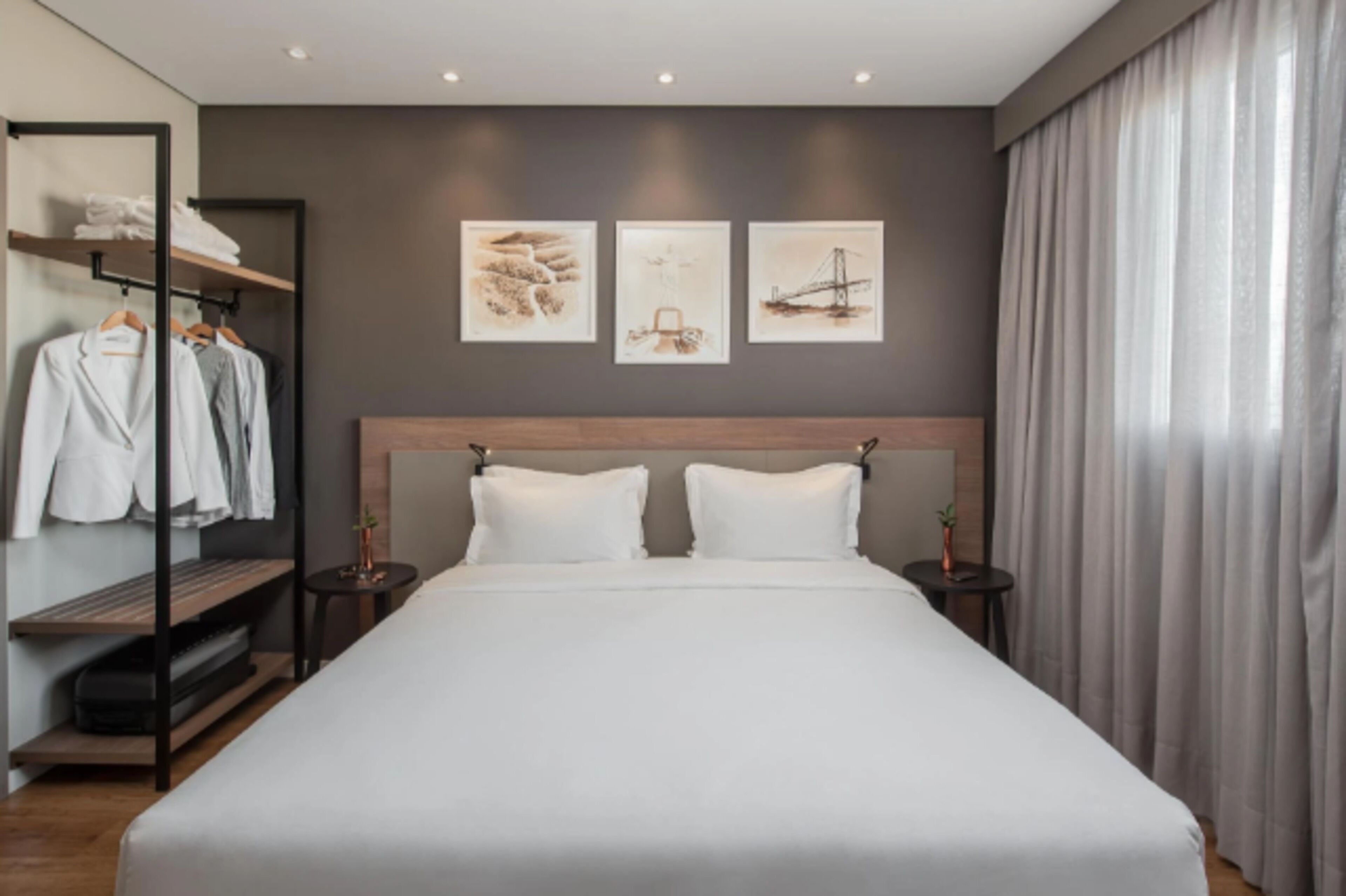 Una habitación de hotel moderna con una cama grande, un armario con ropa colgada y tres cuadros enmarcados en la pared.