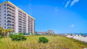 A multi-story hotel with balconies, a beach in the background, and a clear sky.