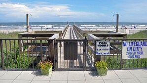 A beachside walkway leading to a pool area with a lifeguard duty sign and a pool hours sign.