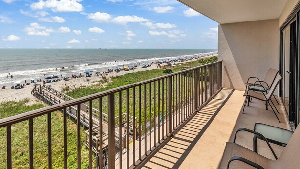 A balcony with a view of the beach, outdoor furniture, and a wooden deck.