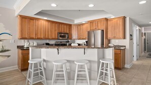 A modern kitchen with wooden cabinets, a stainless steel refrigerator, a microwave, and a granite countertop. There are three white bar stools in front of the counter.