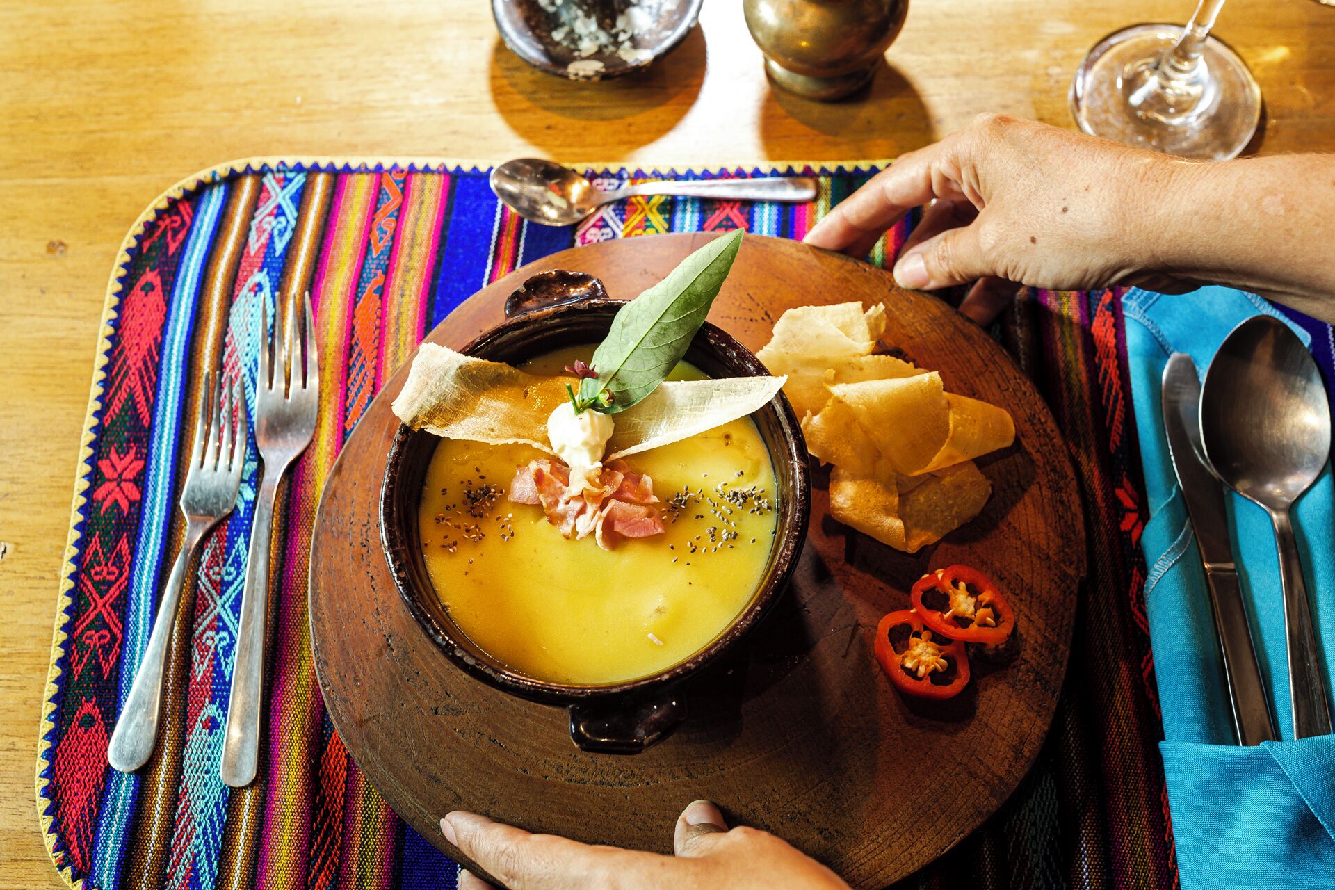 Un tazón de sopa adornado con hierbas y tocino, servido en un plato de madera con totopos y pimiento rojo en rodajas.