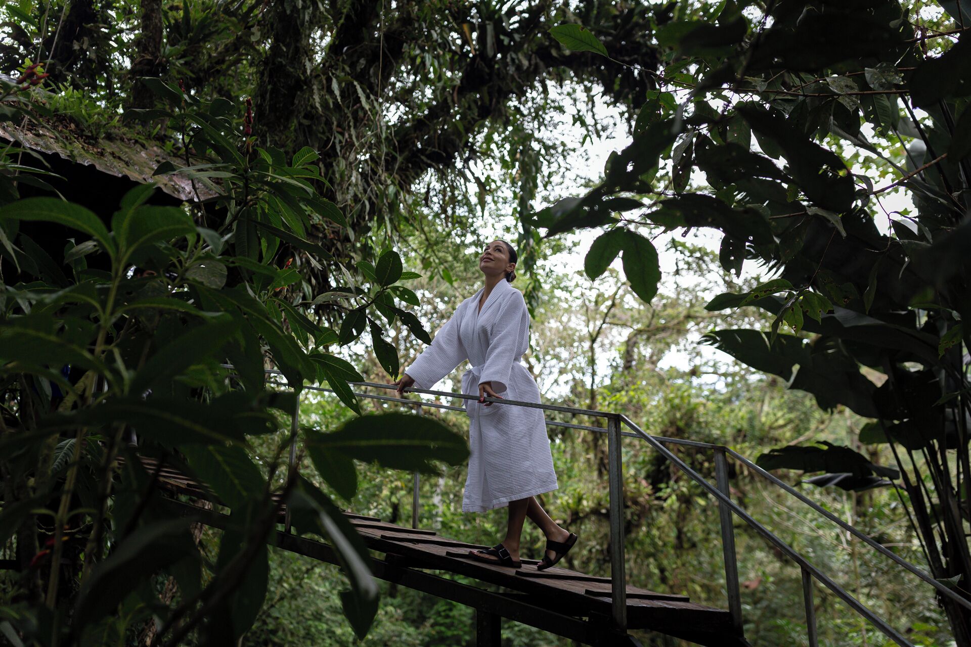 Uma pessoa vestida com um robe branco em uma ponte de madeira, cercada por vegetação densa.