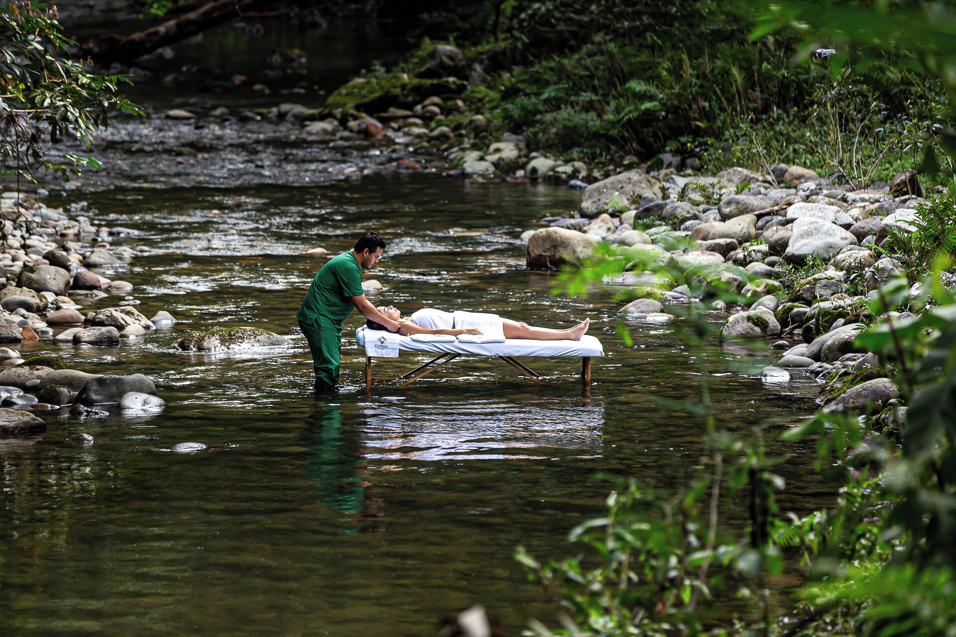 A person lying on a white mat in a natural setting with a river and rocks.
