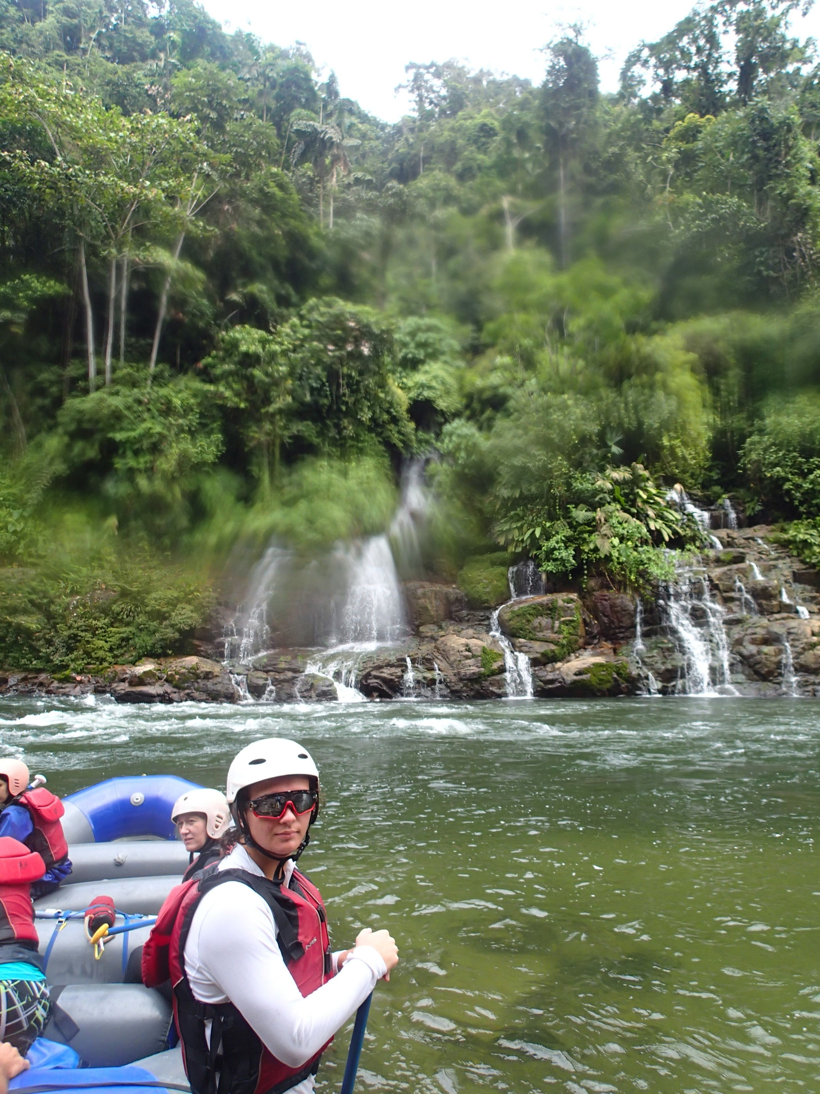 Un groupe de personnes fait du rafting sur une rivière, avec une cascade en arrière-plan.