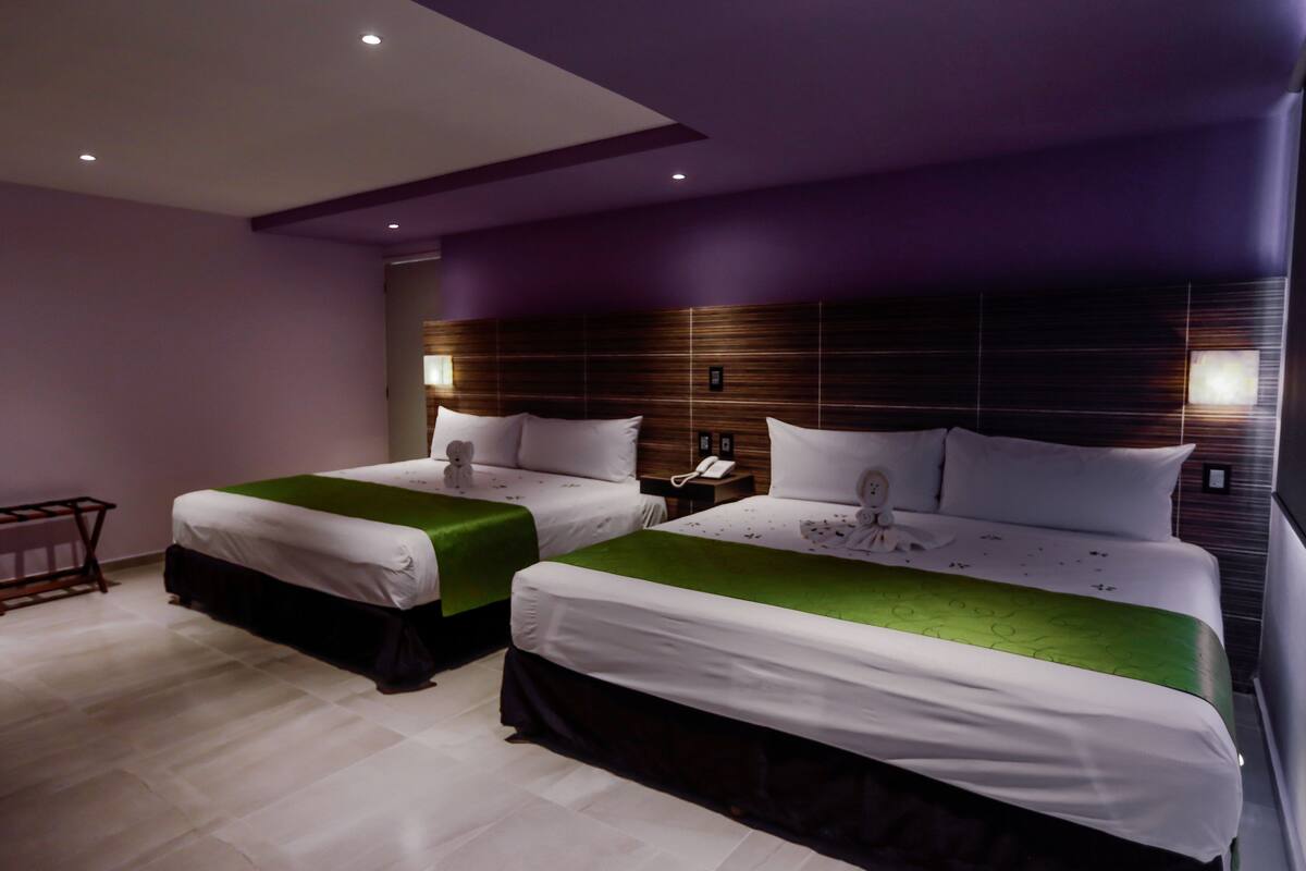 A hotel room with two beds, a wooden headboard, a bedside table with a phone, and a small table with a chair.