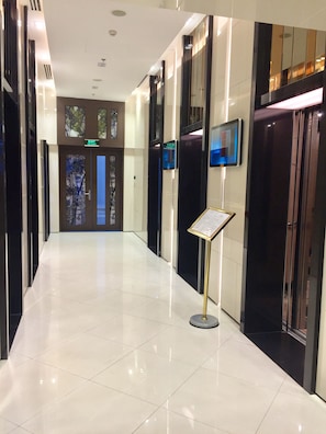 A modern hotel corridor with a shiny tiled floor, glass elevator doors, and a digital information board.
