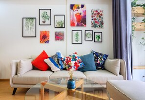 A living room with a sofa, colorful cushions, a glass coffee table with a vase, and framed botanical artwork on the wall.