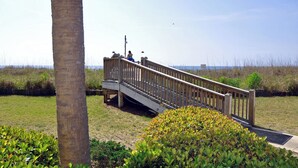 A wooden bridge with railings, a palm tree, and a grassy area with bushes. - Verandas 305- Oceanfront 3BR Condo-Oceanfront Pool- Walk to Main Street (NO. MYRTLE BEACH)