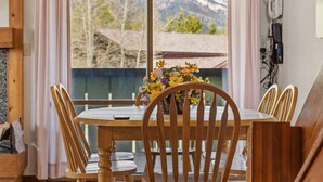 Un comedor con mesa y sillas de madera, un florero con flores y vista a las montañas a través de una ventana.